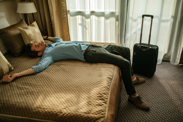 een vermoeide man liggend op een bed door een jetlag in een hotelkamer met een koffer ernaast