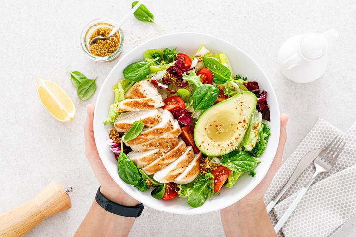 Gegrilde kip en een frisse groentesalade met tomaat, avocado, sla en spinazie. Een gezond en ontgiftend voedingsconcept. Ketogeen dieet. Boeddha-kom in handen op een witte achtergrond, bovenaanzicht. lunchsalades voor onderweg