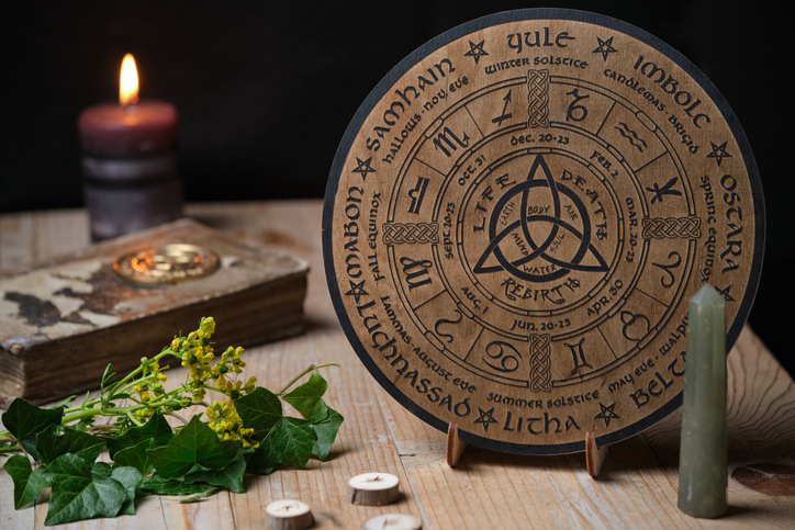 Sfeervol stilleven met een houten Jaarwiel, gegraveerd met de acht heidense feesten. De schijf ligt op een oude houten tafel, omringd door een zwarte rituele kaars, een oud boek, een edelstenen obelisk en runenstenen. De compositie is gefotografeerd in een donkere en stemmige stijl met warme tinten, wat een mystieke, spirituele sfeer oproept. jaarfeesten
