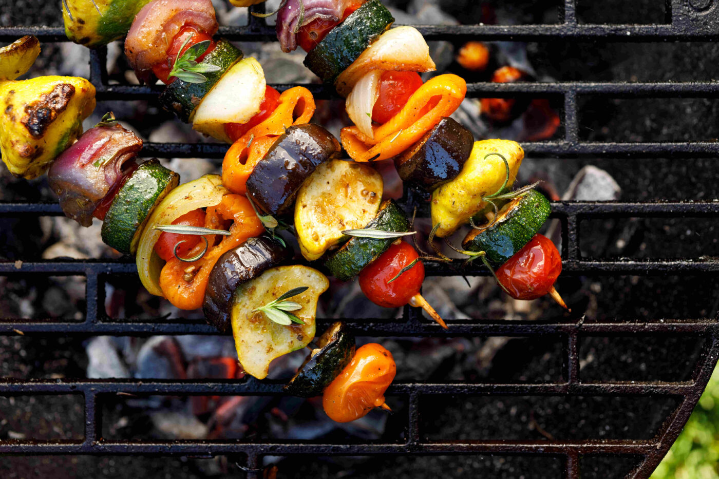 drie spiesjes van courgette, tomaat, paprika, ui, aubergine, besprenkelt met wat rozemarijn, op een barbecue rooster met gloeiende kooltjes eronder